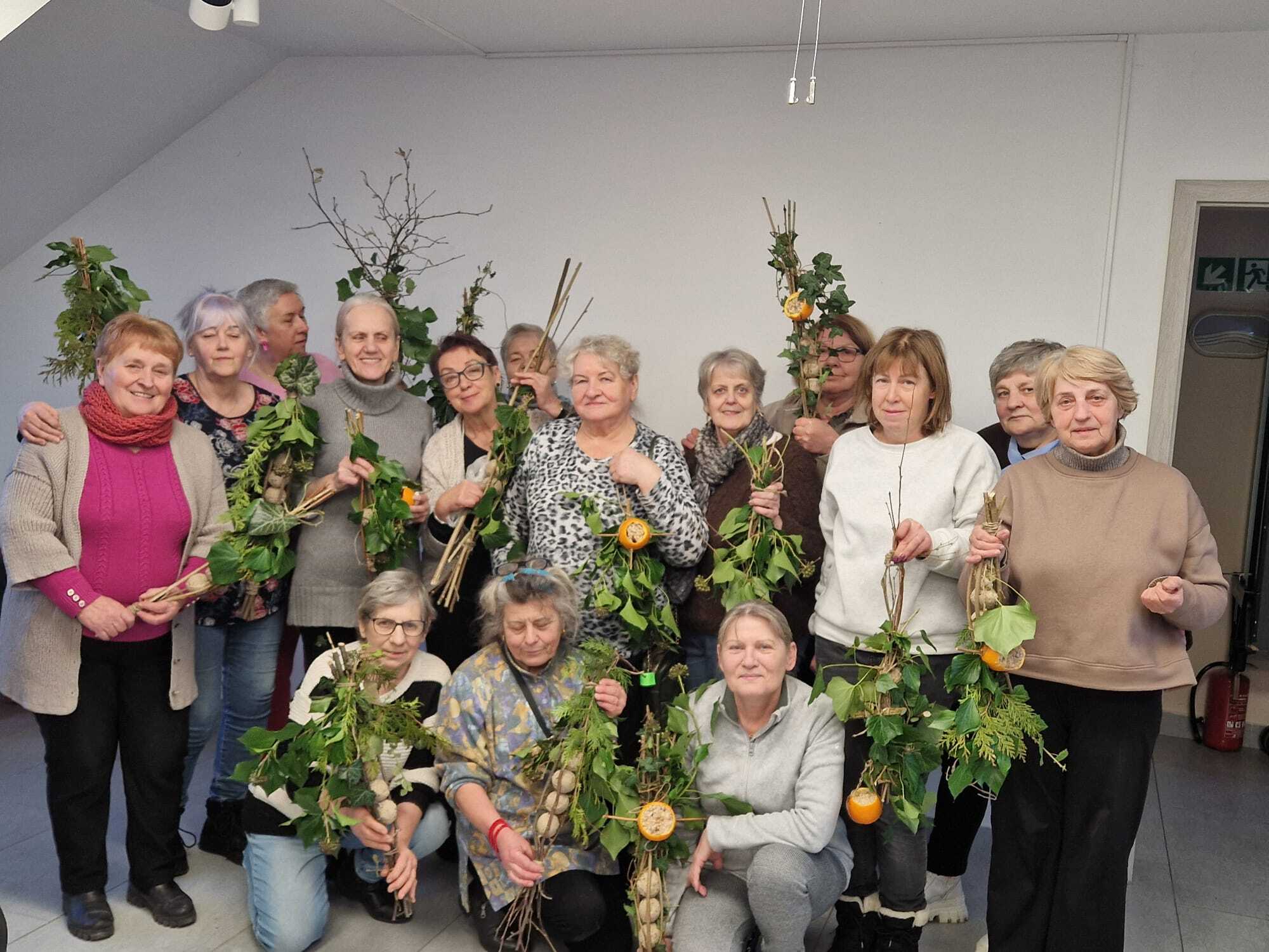 grupa piętnastu uczestniczek warsztatów prezentująca w dłoniach gotowe karmniki z gałązek
