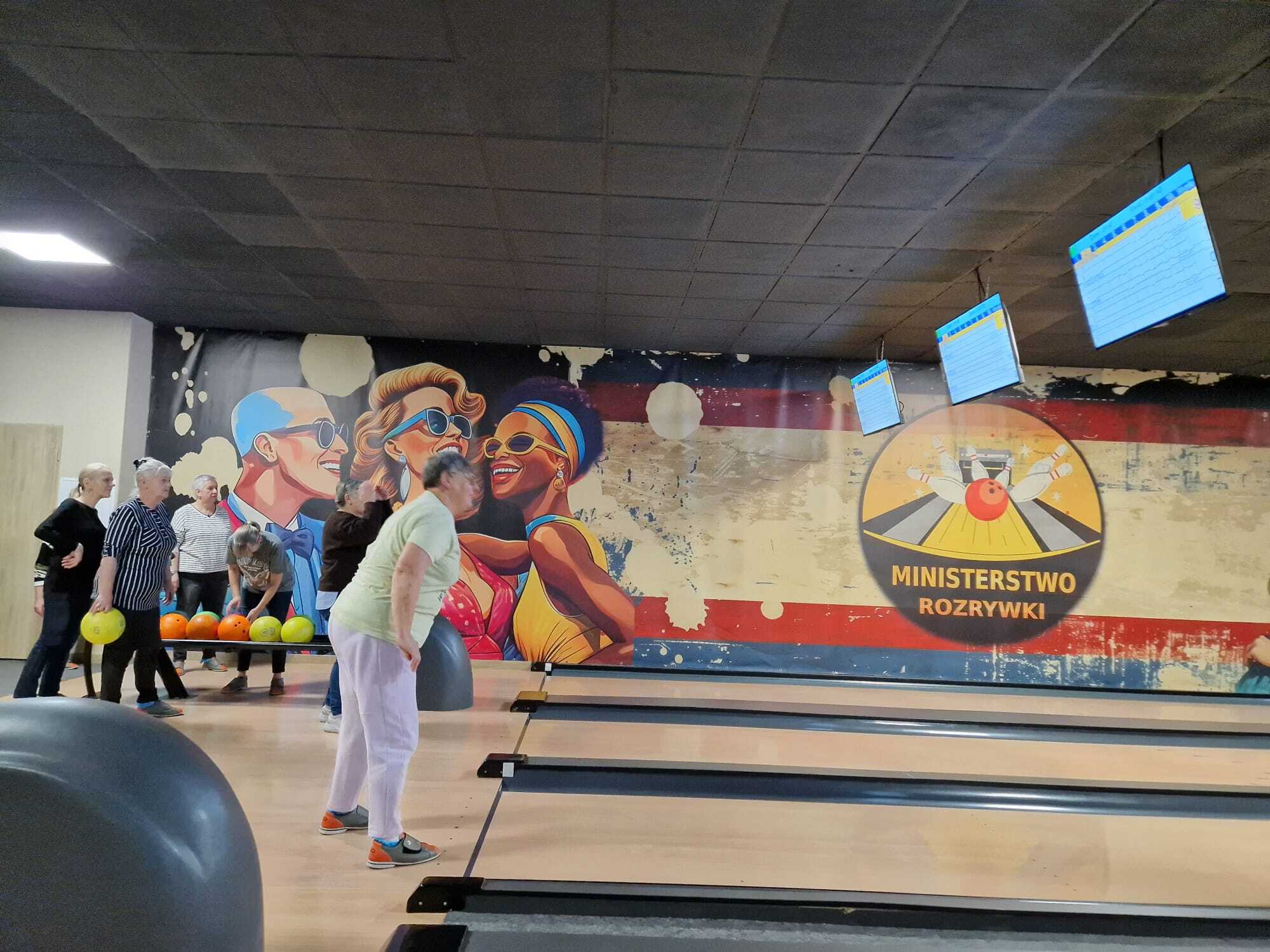 zdjęcie przedstawia uczestniczkę spotkania po oddanym pchnięciu kulą do gry w kręgle, w tle inne uczestniczki szykujące się do swojego zagrania