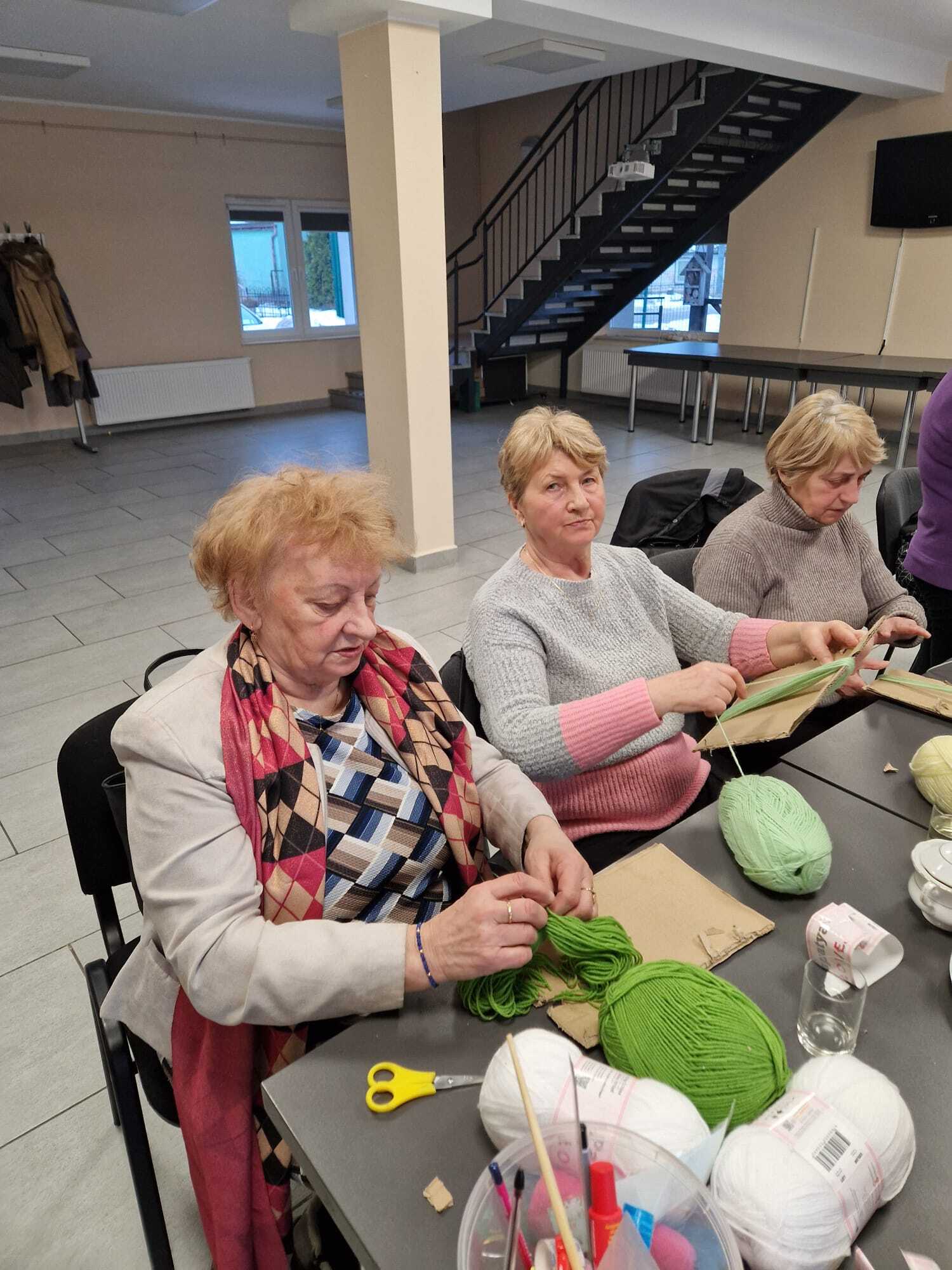 trzy uczestniczki warsztatów siedzące przy stole wykonujące ozdoby z włóczki