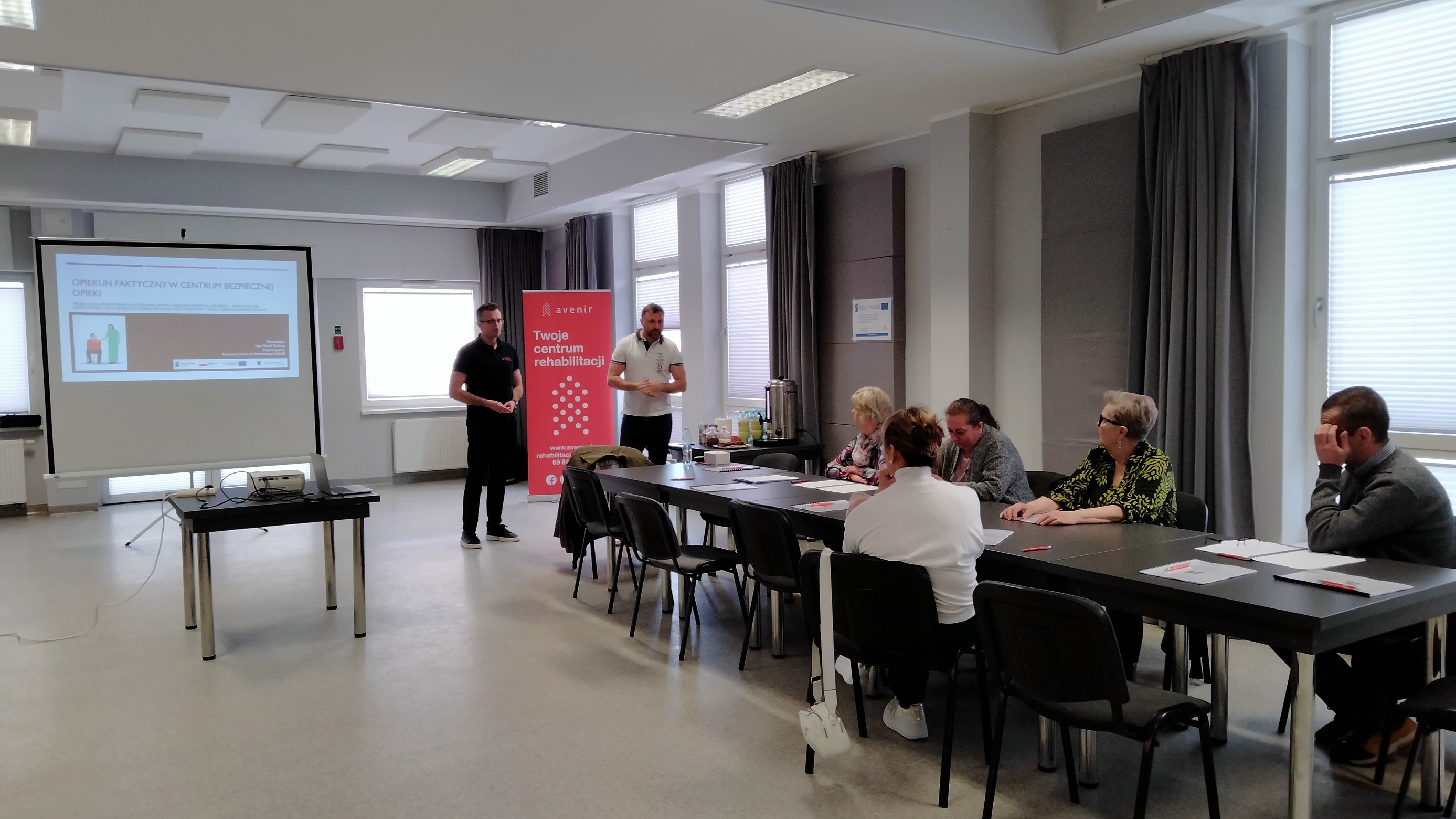 zdjęcie przedstawia uczestników spotkania siedzących przy wspólnym stole słuchających wykładu dwóch prowadzących stojących przy ekranie projekcyjnym