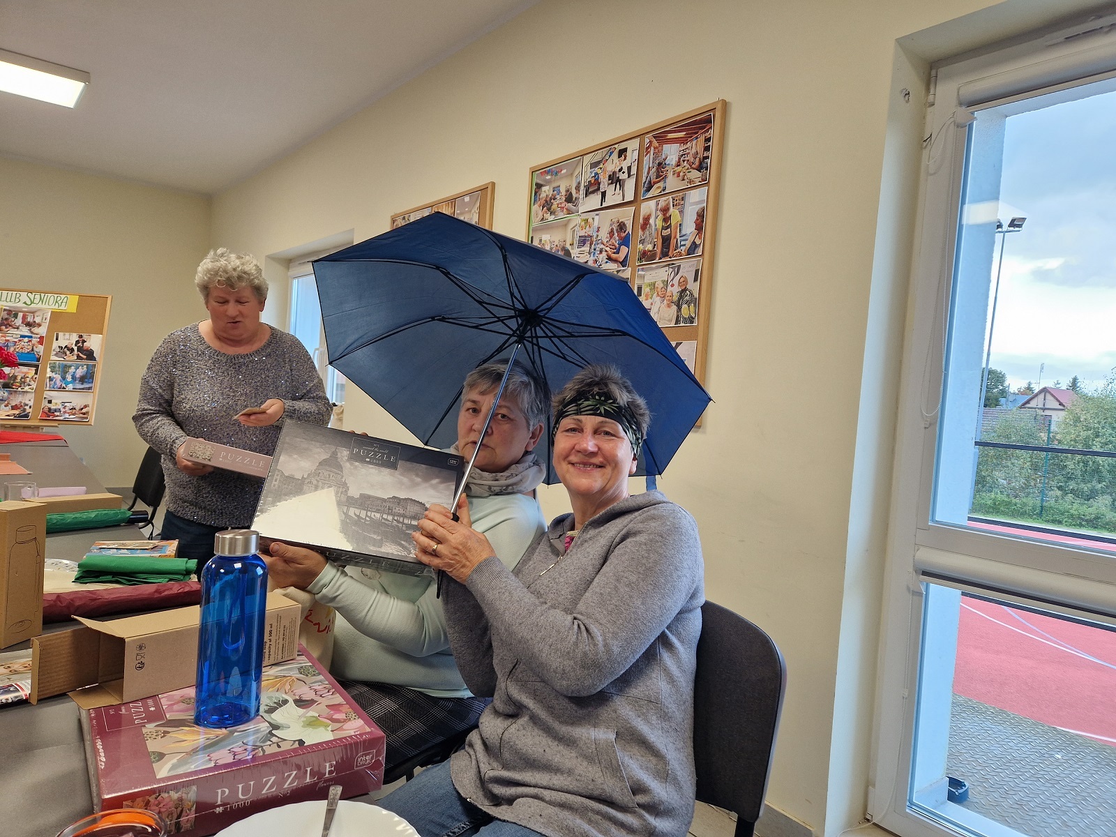 dwie uczestniczki spotkania siedzące przy stole trzymające w rękach upominki: parasol i puzzle