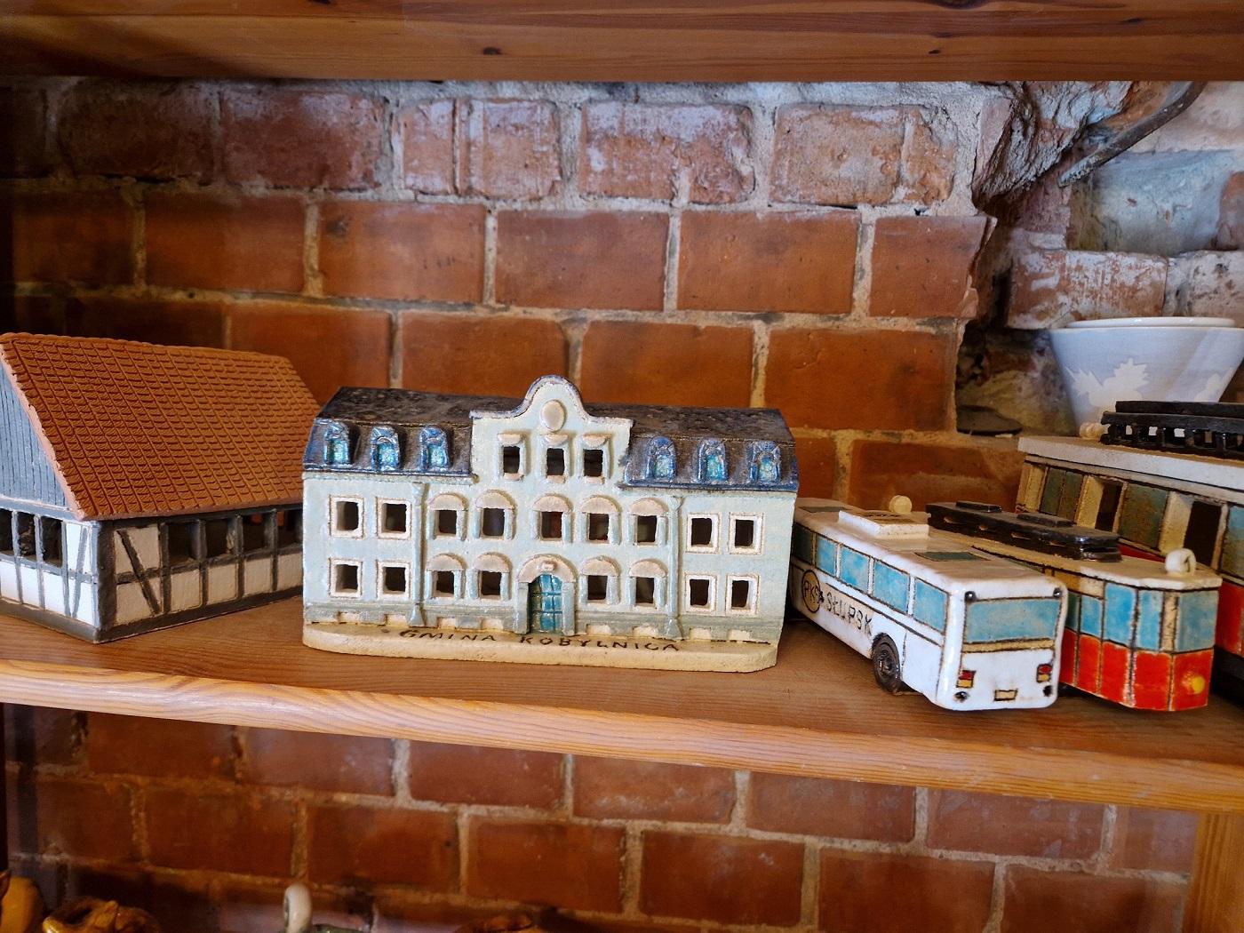 gotowe wyroby ceramiczne umieszczone na półce ceramikarni przedstawiające dwa budynki, biały autobus i czerwony tramwaj